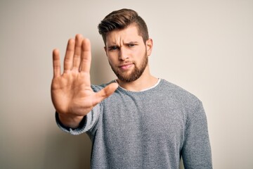 Young handsome blond man with beard and blue eyes wearing casual sweater doing stop sing with palm...