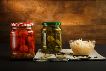 Jars with pickled tomatoes and cucumbers and sauerkraut on a wooden table on a black background.