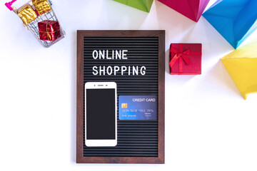 Miniature of gift boxes in trolley, smartphone, word on black frame, credit card and colorful bags on white desk. Online shopping, technology and lifestyle concept. Top view, flat lay and copy space