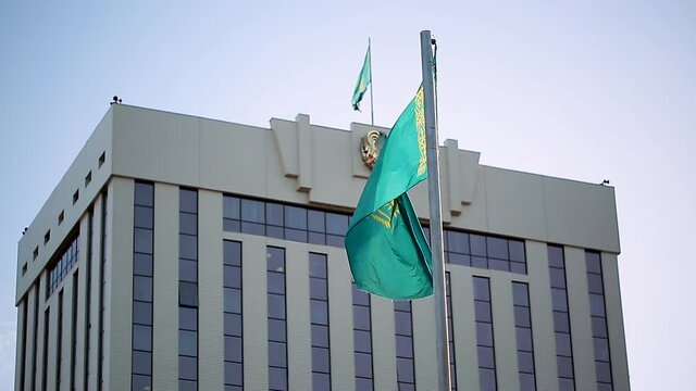 The flag of Kazakhstan is developing in the wind against the background of a government building with a coat of arms. The city of Nur Sultan is the administrative center