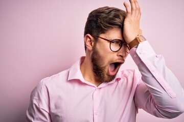 Young handsome blond man with beard and blue eyes wearing pink shirt and glasses surprised with...