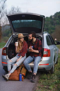 Couple sit on the trunk of car and wonder at photo on tablet. Girl smile and drink form eco cup. Time to Travel concept