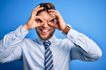Young blond businessman with beard and blue eyes wearing elegant shirt and tie standing doing ok gesture like binoculars sticking tongue out, eyes looking through fingers. Crazy expression.