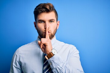 Young blond businessman with beard and blue eyes wearing elegant shirt and tie standing asking to be quiet with finger on lips. Silence and secret concept.