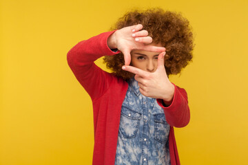 Portrait of woman with fluffy curly hair focusing eye at camera through photo frame gesture, cropping view, capturing moment, concentrating at target. indoor studio shot isolated on yellow background