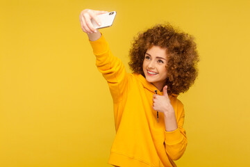 Portrait of optimistic trendy curly-haired woman in urban style hoodie taking selfie, having conversation on video call and showing thumbs up, like gesture. studio shot isolated on yellow background