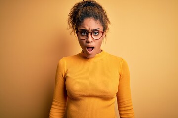 Young beautiful african american girl wearing sweater and glasses over yellow background afraid and shocked with surprise and amazed expression, fear and excited face.