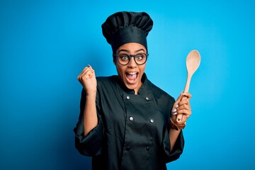 Young african american chef girl wearing cooker uniform and hat holding wooden spoon tool screaming...