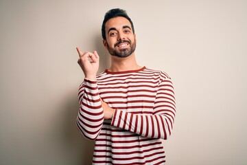 Fototapeta premium Young handsome man with beard wearing casual striped t-shirt standing over white background with a big smile on face, pointing with hand and finger to the side looking at the camera.
