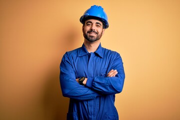 Mechanic man with beard wearing blue uniform and safety helmet over yellow background happy face...