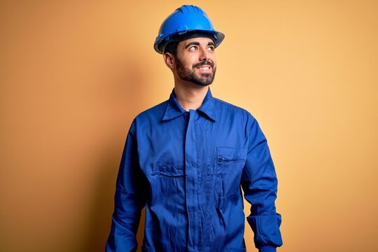 Mechanic man with beard wearing blue uniform and safety helmet over yellow background looking away to side with smile on face, natural expression. Laughing confident.