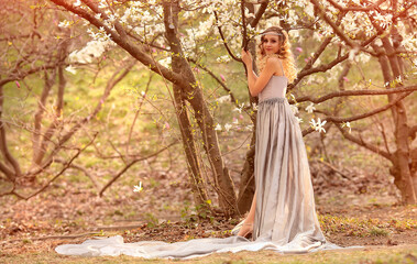 Obraz na płótnie Canvas Young beauty blonde with jewelry for hair, in a silver dress with a long train in the garden where magnolia trees bloom. Beauty and fashion concept. Flares, bokeh effects.Copy space on the left