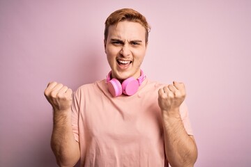 Young handsome redhead man listening to music using headphones over pink background celebrating surprised and amazed for success with arms raised and open eyes. Winner concept.