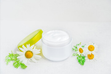 Obraz na płótnie Canvas Herbal cosmetic body cream with chamomile oil in opened plastic container and fresh flowers on white background. Natural organic moisturizer and cleansing skincare product. Selective focus. Copy space