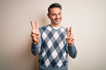 Young handsome man wearing casual sweater standing over isolated white background smiling looking to the camera showing fingers doing victory sign. Number two.