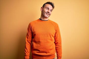 Young handsome man wearing orange casual sweater standing over isolated yellow background looking away to side with smile on face, natural expression. Laughing confident.