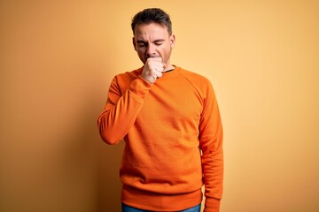 Young handsome man wearing orange casual sweater standing over isolated yellow background feeling unwell and coughing as symptom for cold or bronchitis. Health care concept.