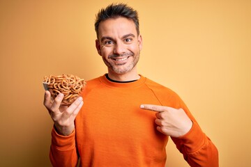 Young handsome man holding bowl with german baked pretzels over yellow background with surprise...