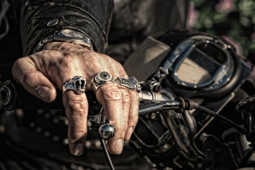A biker's hand, wearing several rings, rests on the handlebar of his motorcycle, with a vintage helmet and goggles in the background