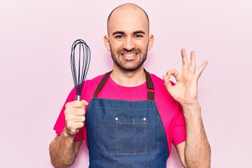 Young handsome bald man wearing apron holding whisk doing ok sign with fingers, smiling friendly gesturing excellent symbol