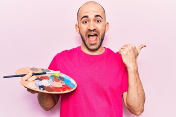 Young handsome bald man painting using paintbrush and palette pointing thumb up to the side smiling happy with open mouth