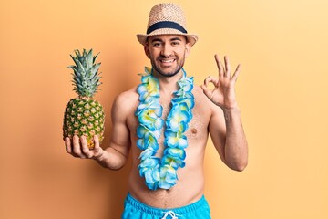 Young handsome bald man wearing swimwear and hawaiian lei holding pineapple doing ok sign with...