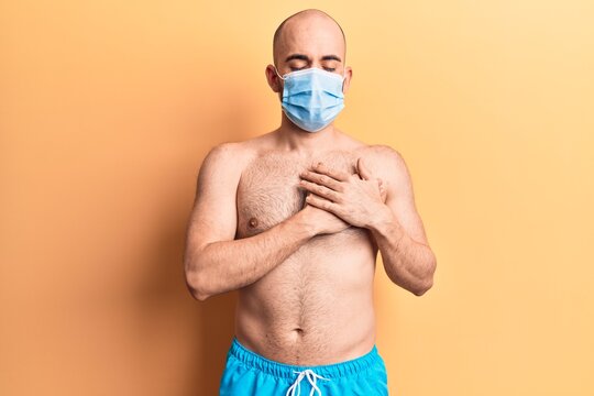Young handsome bald man shirtless wearing swimwear and medical mask smiling with hands on chest, eyes closed with grateful gesture on face. health concept.