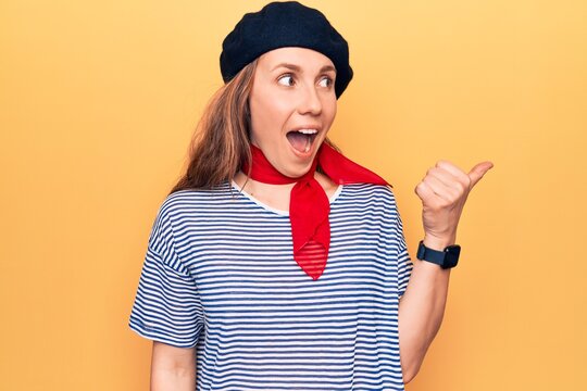 Young beautiful blonde woman wearing french fashion style with scarf and beret pointing thumb up to the side smiling happy with open mouth