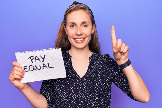 Young beautiful blonde woman holding paper with pay equal protest text smiling with an idea or question pointing finger with happy face, number one