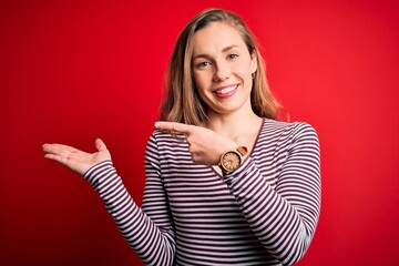 Young beautiful blonde woman wearing casual striped t-shirt over isolated red background amazed and smiling to the camera while presenting with hand and pointing with finger.