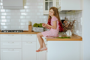 Young cute long haired barefooted girl in stripped summer dress sitting on a kitchen table and holding cherries.