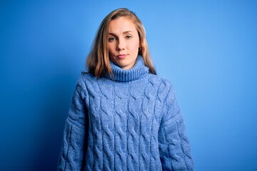 Young beautiful blonde woman wearing casual turtleneck sweater over blue background Relaxed with serious expression on face. Simple and natural looking at the camera.