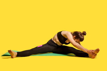 Slim gymnast woman with hair bun in tight sportswear sitting on mat with spread legs, bending to...
