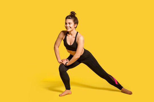 Happy athletic girl with hair bun in tight sportswear doing lower body sport exercise, stretching legs, warming up training muscles for flexibility. indoor full length studio shot, isolated on yellow