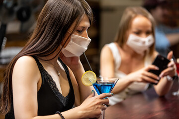 girls in medical masks chatting in a pub. girls in medical masks are talking at the bar. girls in medical masks drink cocktails