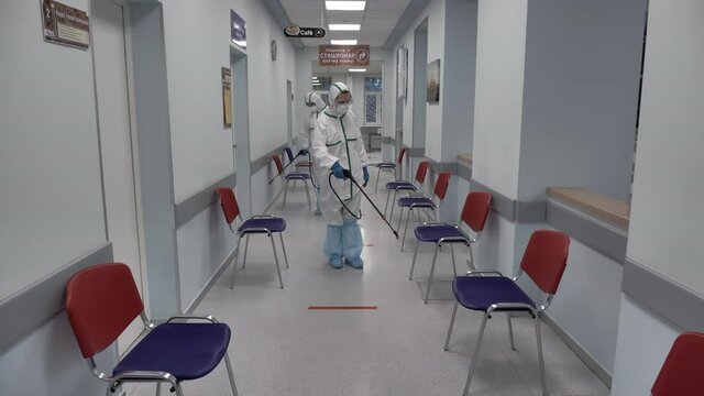 Two Workers In Protective White Suits In Masks, Gloves, Glasses And Shoe Covers Walk Along The Hospital Corridor And Carry Out Sanitization By Spraying A Disinfectant Solution.