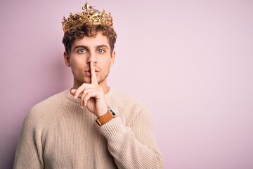 Young blond man with curly hair wearing golden crown of king over pink background asking to be quiet with finger on lips. Silence and secret concept.