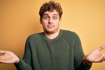 Young blond handsome man with curly hair wearing green sweater over yellow background clueless and...