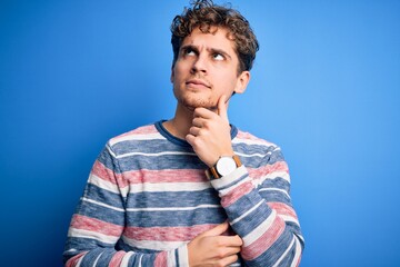 Young blond handsome man with curly hair wearing striped sweater over blue background with hand on chin thinking about question, pensive expression. Smiling with thoughtful face. Doubt concept.