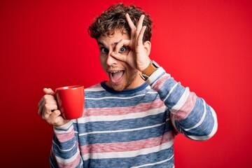 Young blond man with curly hair drinking cup of coffee standing over red background with happy face smiling doing ok sign with hand on eye looking through fingers