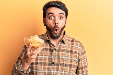 Young hispanic man holding potato chip scared and amazed with open mouth for surprise, disbelief...
