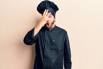 Young hispanic man wearing cooker uniform peeking in shock covering face and eyes with hand,...
