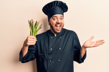 Young hispanic man wearing cooker uniform holding asparagus celebrating achievement with happy...