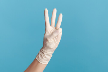 Profile side view closeup of human hand in white surgical gloves showing number 3 three with hands. indoor, studio shot, isolated on blue background.