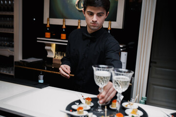 portrait of a young man in a restaurant. the waiter in a black shirt put the glasses on the tray.