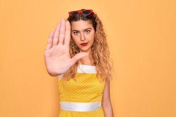 Beautiful blonde woman pin-up with blue eyes wearing red sunglasses over yellow background with open hand doing stop sign with serious and confident expression, defense gesture