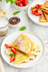 Thin pancakes or crepes with chocolate cream, banana and strawberries on a white plate with a glass of cappuccino on a light wooden background. Vertical orientation. Copy space.