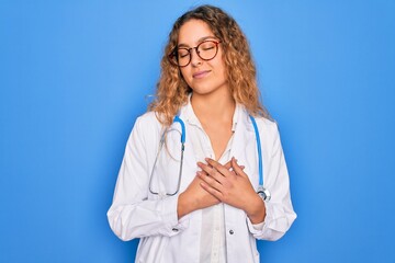 Young beautiful blonde doctor woman with blue eyes wearing coat and stethoscope smiling with hands on chest with closed eyes and grateful gesture on face. Health concept.