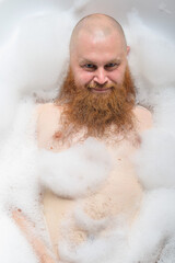 Portrait of a bald man with a long red beard takes a bath with foam. Top view on a cheerful funny guy in soapy water.