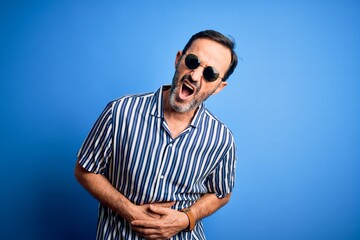 Middle age hoary man wearing striped shirt and sunglasses over isolated blue background with hand on stomach because nausea, painful disease feeling unwell. Ache concept.